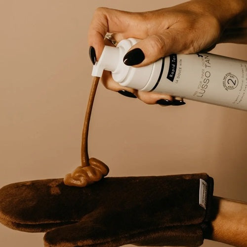 Hand applying Lusso Rapid Express Self Tan Mousse from a bottle labelled 'Lusso Tan' onto a brown glove against a beige background.