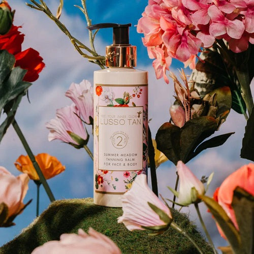 Lusso Tan summer meadows tanning balm surrounded by flowers with a blurred sky background
